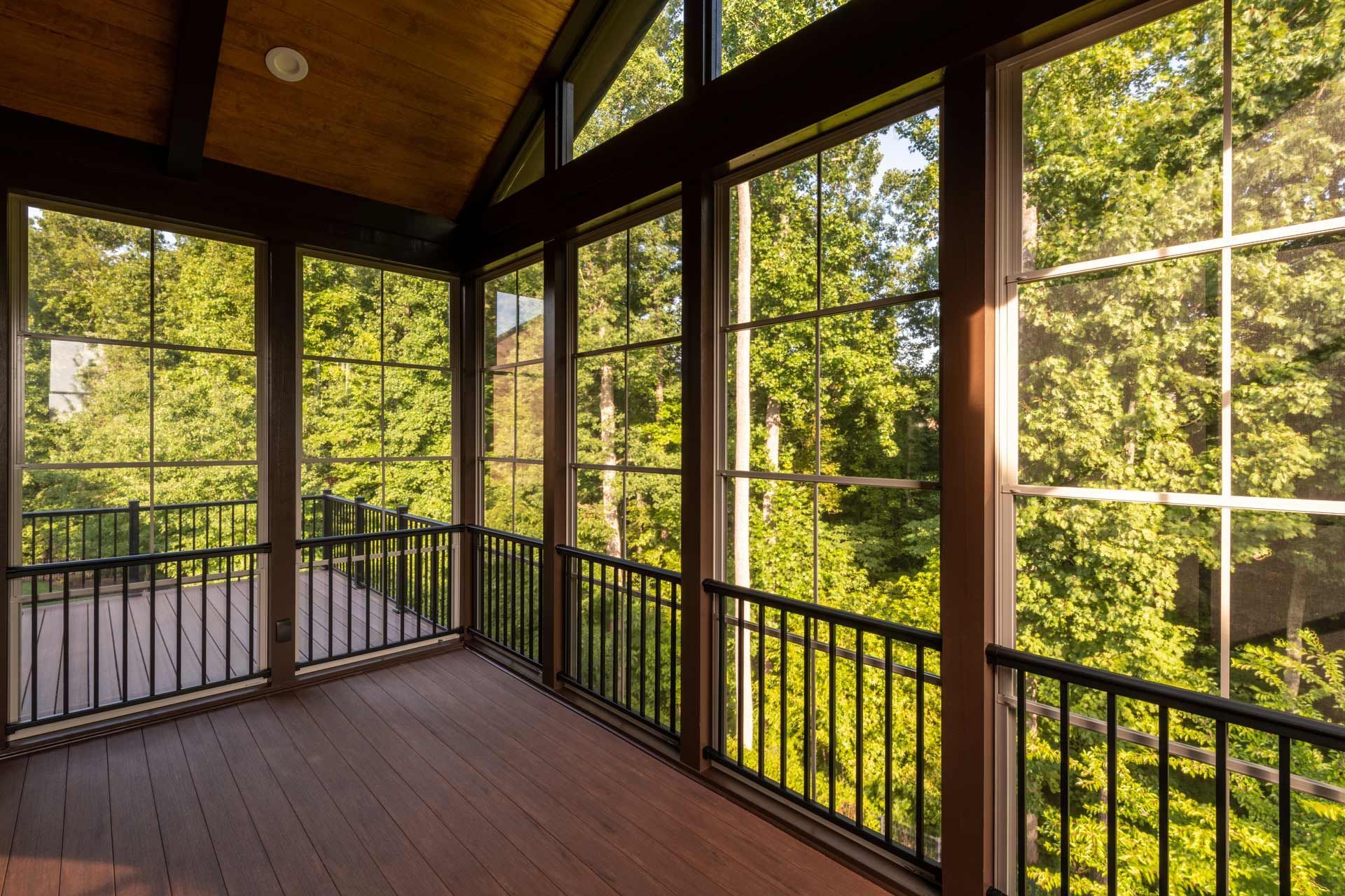 Sunlit screened-in porch overlooking trees; brown deck, railings, and window frames; green foliage in background.