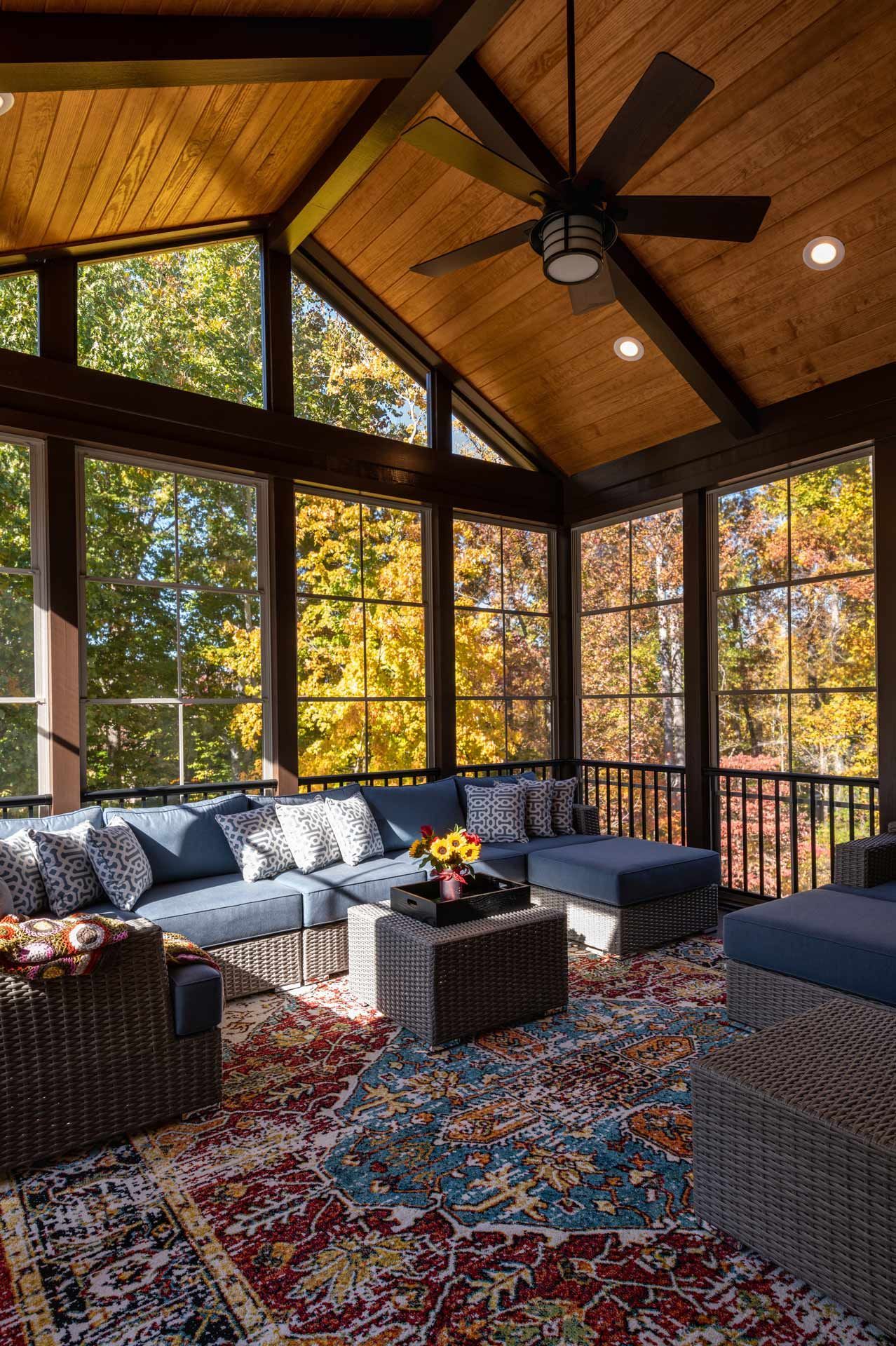 Screened-in porch with blue furniture, area rug, and ceiling fan, surrounded by fall foliage.