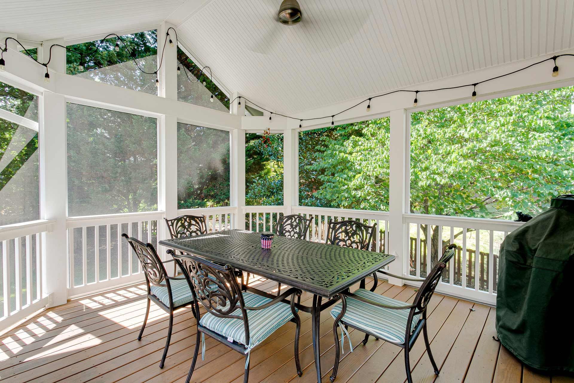 Screened-in porch with a metal table and chairs, surrounded by trees. String lights and a ceiling fan are visible.