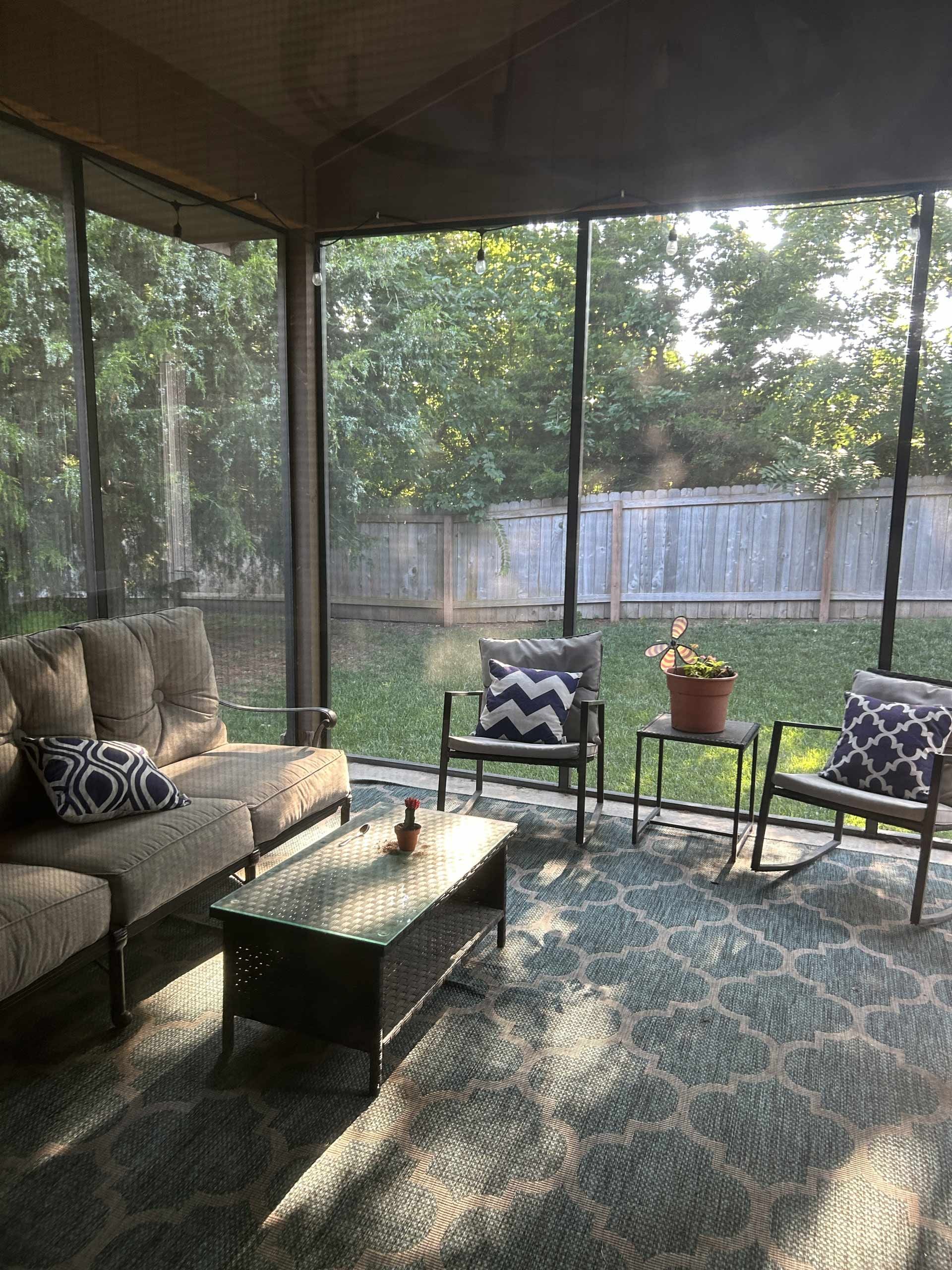 Screened-in porch with a couch, chairs, rug, and a coffee table facing a backyard with trees and a fence.