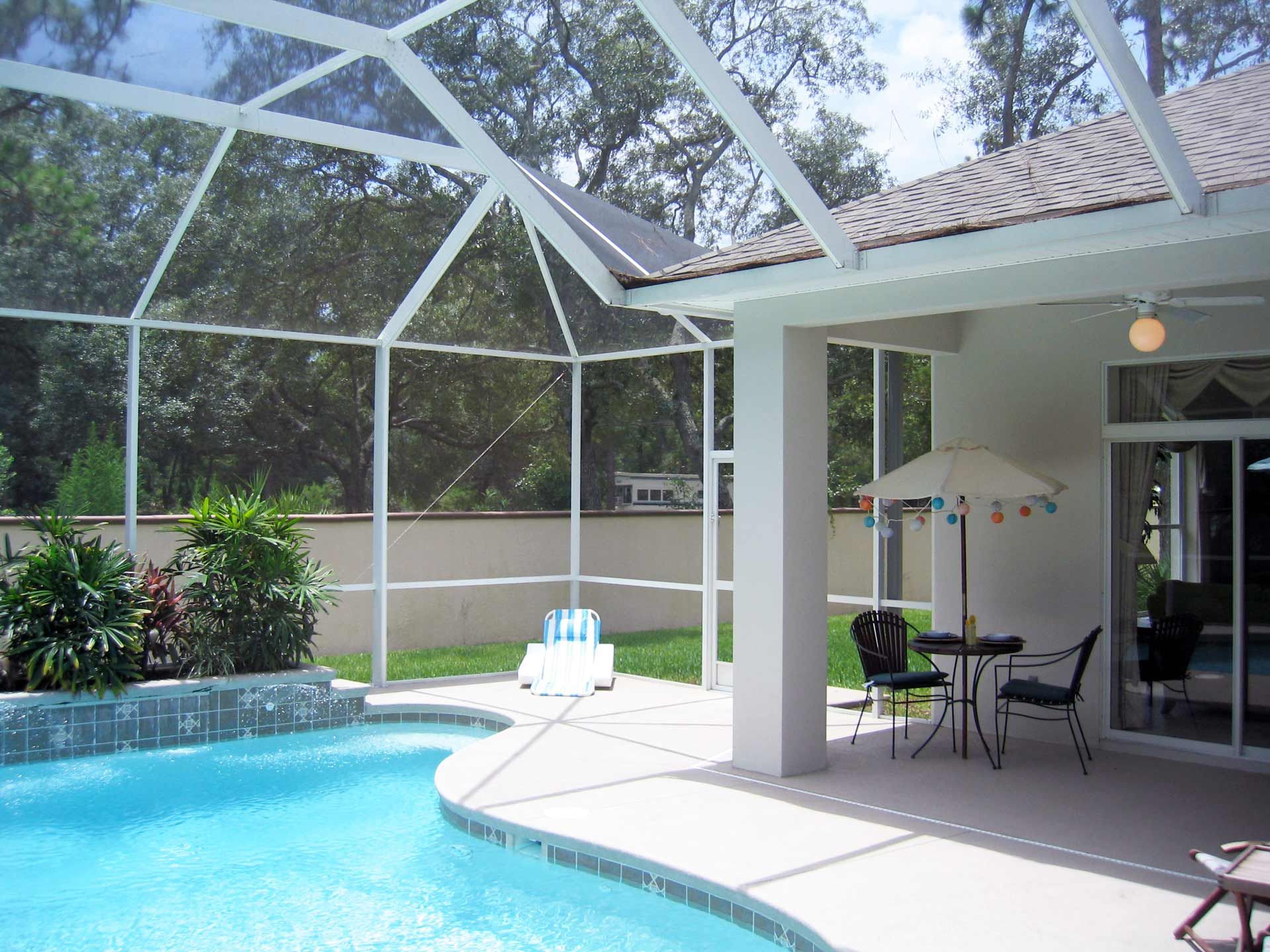 A screened-in pool area with a pool, patio furniture, and white framing against a house and trees.