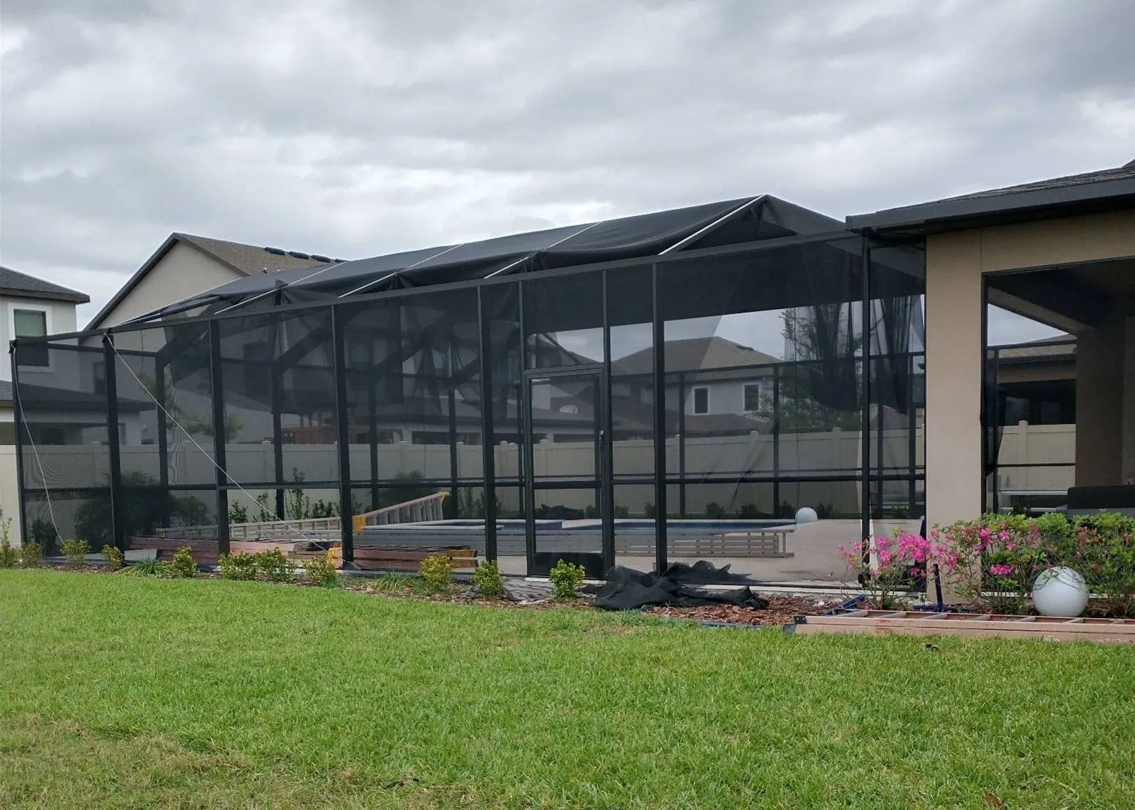 Pool enclosure with a screened roof, framed by a green lawn and residential buildings under an overcast sky.