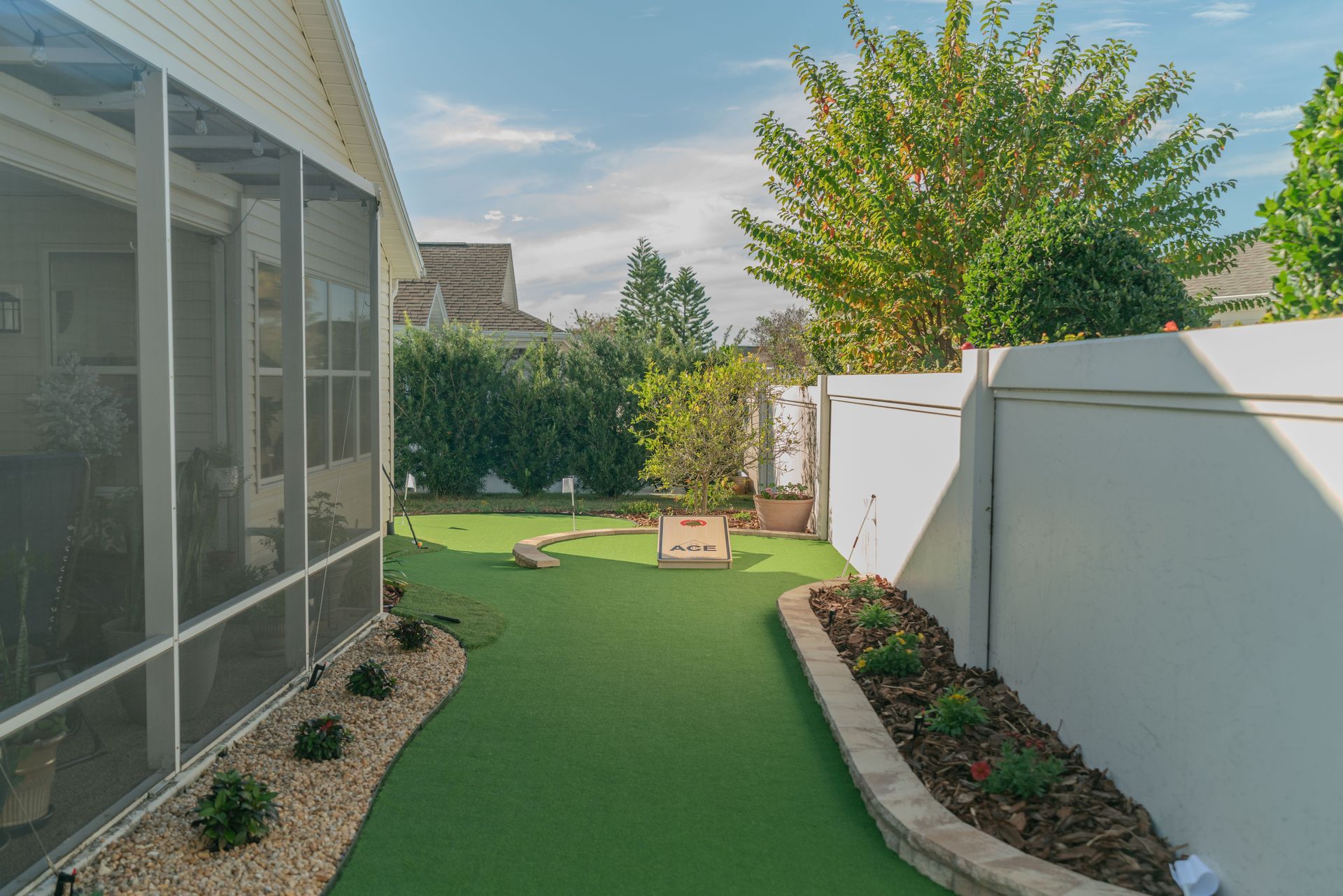 Backyard with green turf, white fence, and screened porch. Landscaping with shrubs and a tree. Sunny day.
