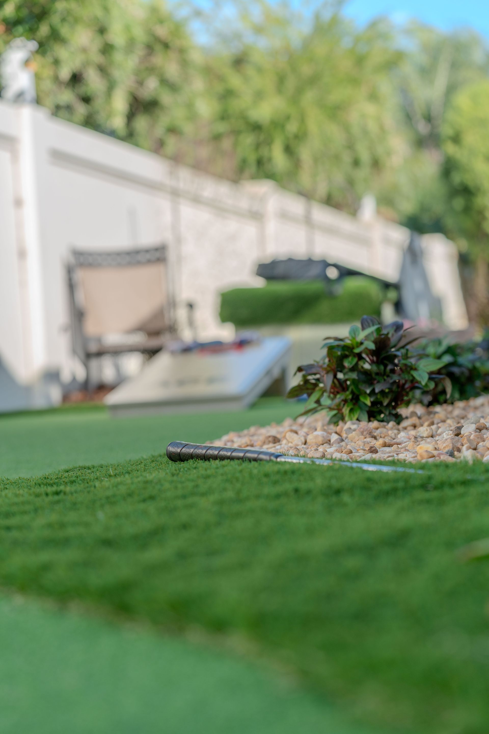 Green lawn with a gravel bed and greenery in the foreground, white wall and trees blurred in background.
