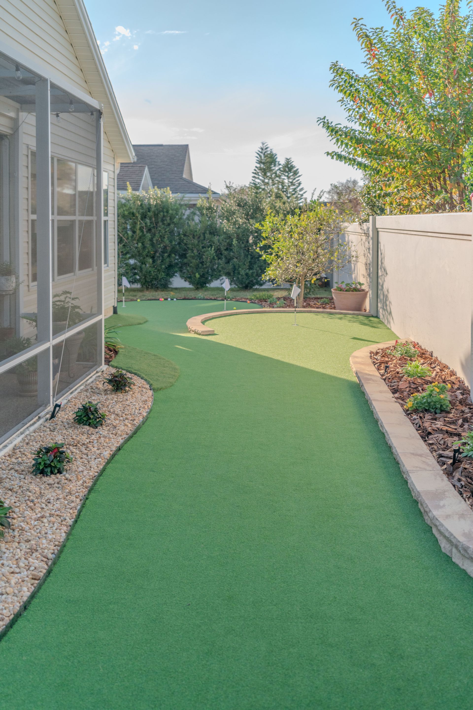 Green artificial lawn with stone edging, screen porch, and garden area with trees.