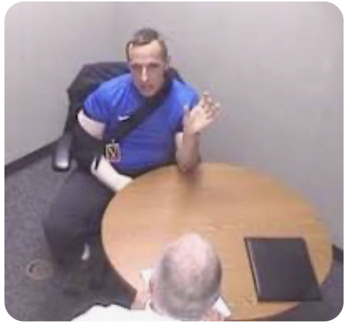 A man in a blue shirt is sitting at a table talking to another man