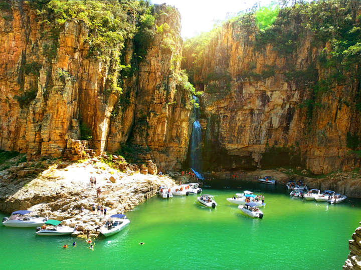 Lagoa turquesa com pequenos barcos sob altas falésias alaranjadas e árvores verdes.
