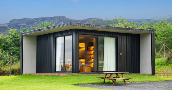 Cabana moderna e compacta com revestimento preto, interior iluminado e mesa de piquenique em um gramado com vista para as montanhas.