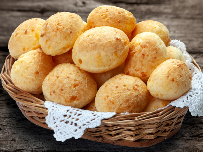 Cesta de pãezinhos dourados assados, coberta com uma toalha de renda, sobre uma mesa de madeira.