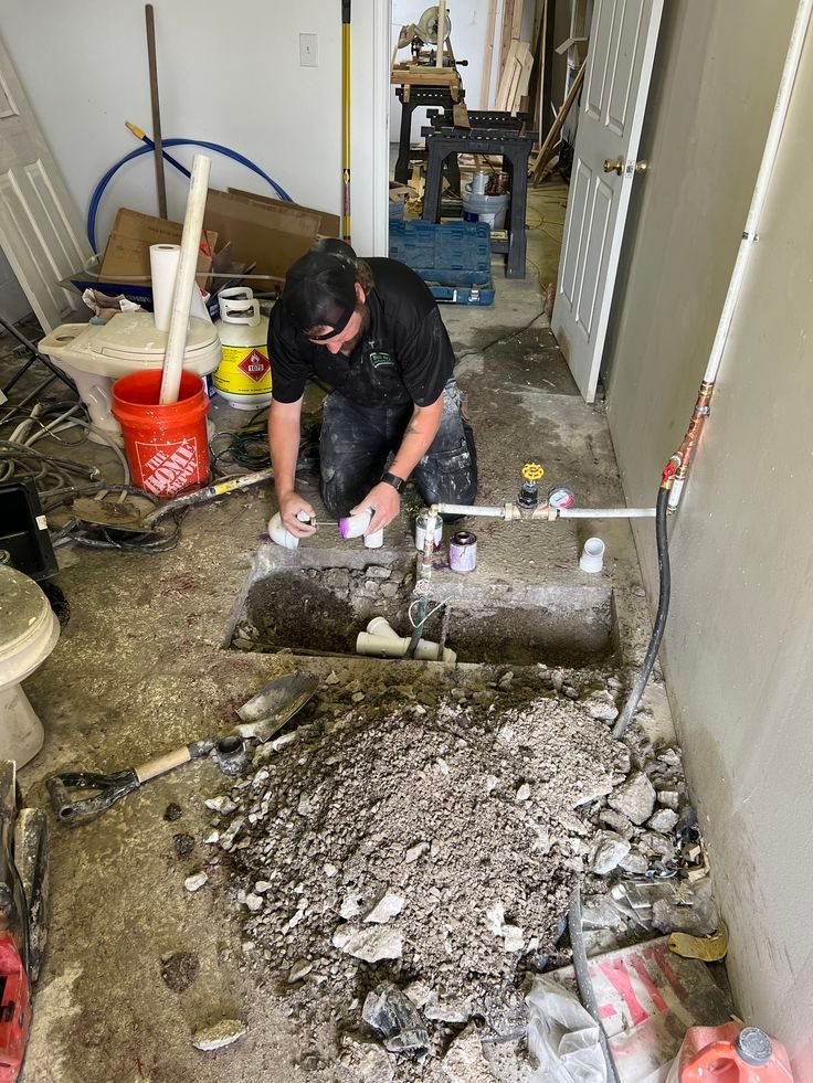 A worker kneels on a concrete floor, repairing plumbing pipes inside a dug-out trench in a room under renovation.