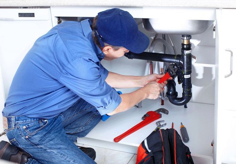 A plumber in a blue uniform kneels under a kitchen sink, using a tool to repair the dark plastic piping.