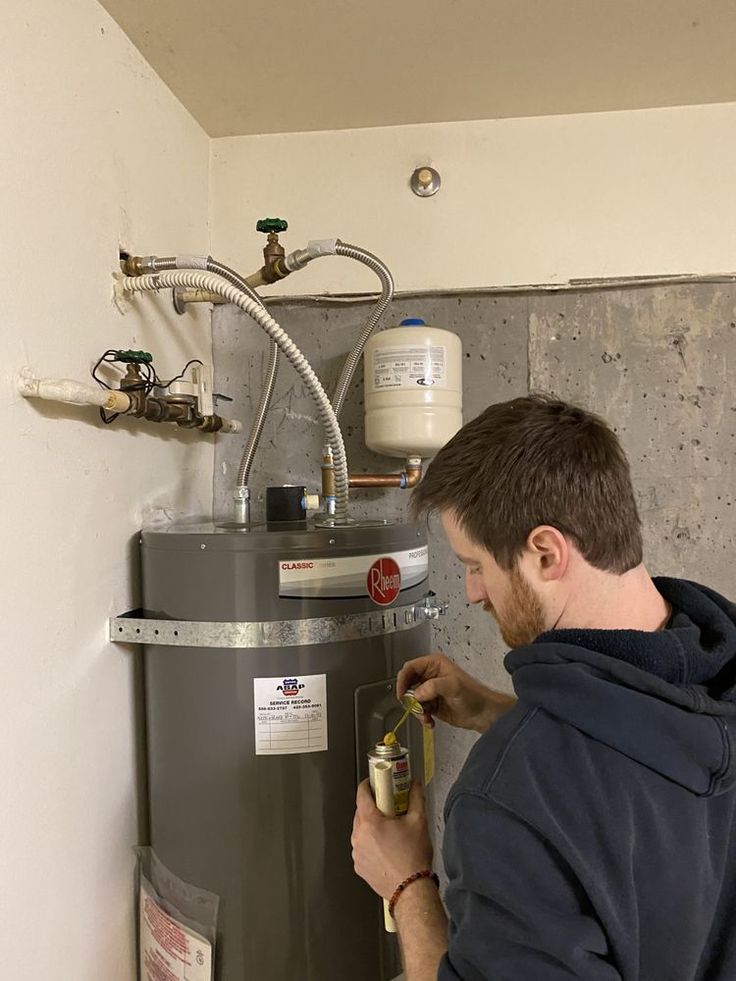 A person performing maintenance on a gray water heater in a utility room, working near the pipes and expansion tank.