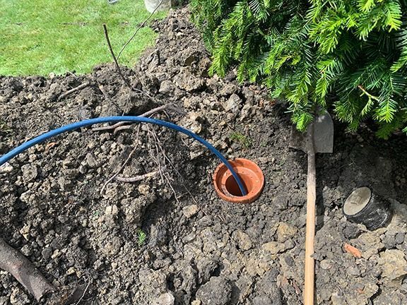 A blue irrigation pipe runs into a terracotta pipe fitting set in the ground next to a shovel and evergreen shrub.