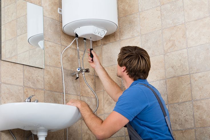 A technician in a blue shirt repairs a wall-mounted electric water heater above a bathroom sink.