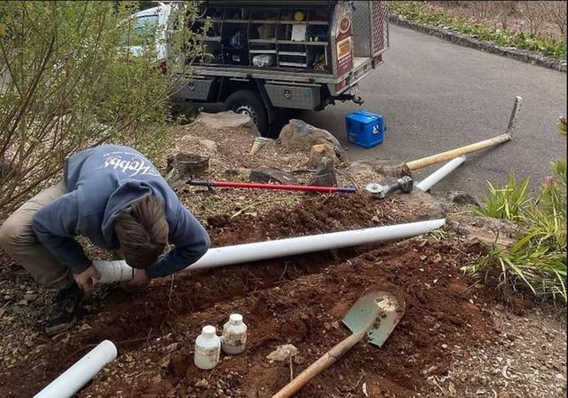 A person in a blue hoodie kneels in a dirt trench outdoors, working on connecting white PVC drainage pipes.