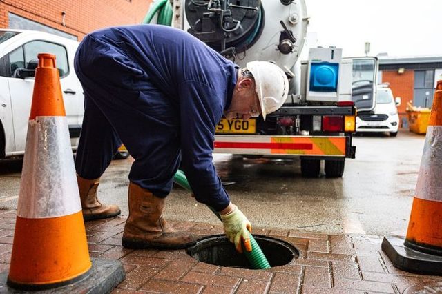 A utility worker in protective gear feeds a green suction hose into an open manhole, flanked by orange traffic cones.