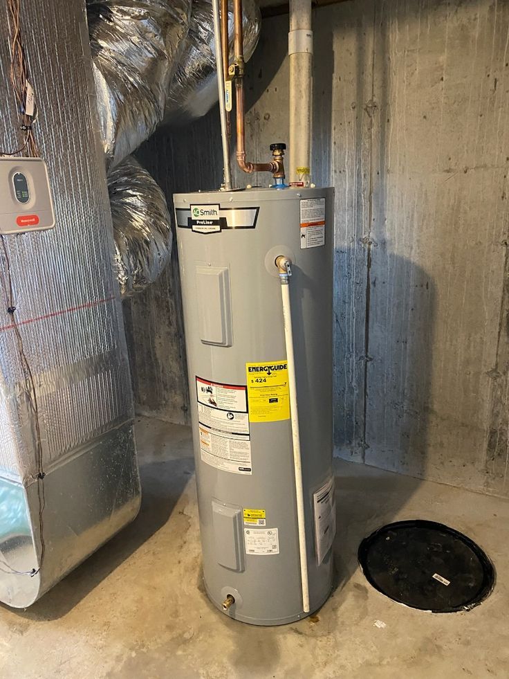 A grey water heater sits in a basement next to metallic ductwork and a black drain cover on a concrete floor.