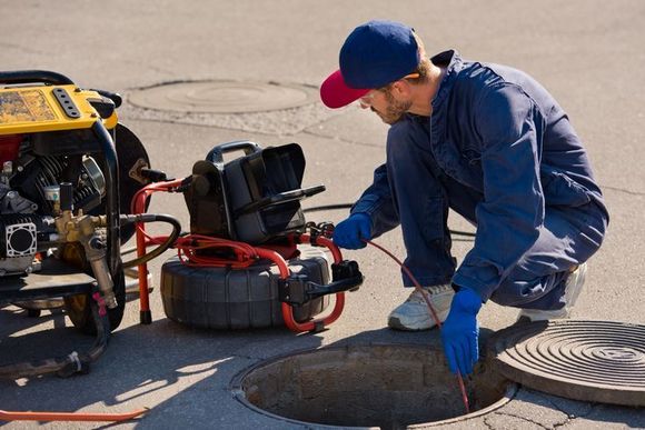 A worker in a blue uniform kneels by an open manhole, operating a camera inspection system on the paved ground.