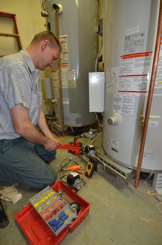 A person kneels while using a red pipe wrench on gas lines near two large water heaters in a utility room.