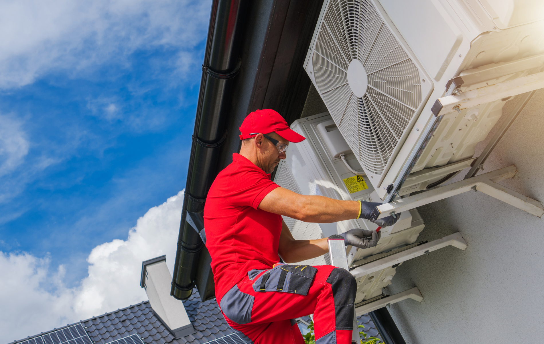 A technician in a red uniform installs or repairs an outdoor air conditioning unit mounted on a building wall.