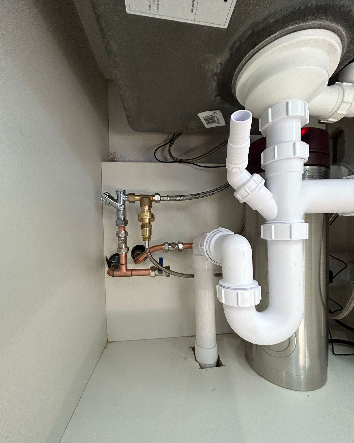 Under-sink plumbing showing a white P-trap, copper pipes, a brass valve, and an open drainage hose connector.