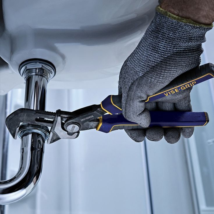 A gloved hand uses blue and yellow VISE-GRIP pliers to tighten a chrome pipe connection under a bathroom sink.