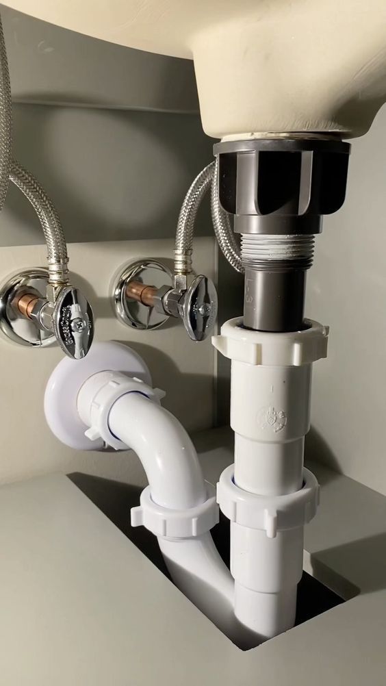 A bathroom sink drain assembly under a basin, featuring white PVC pipes and metal water supply lines against a grey cabinet.
