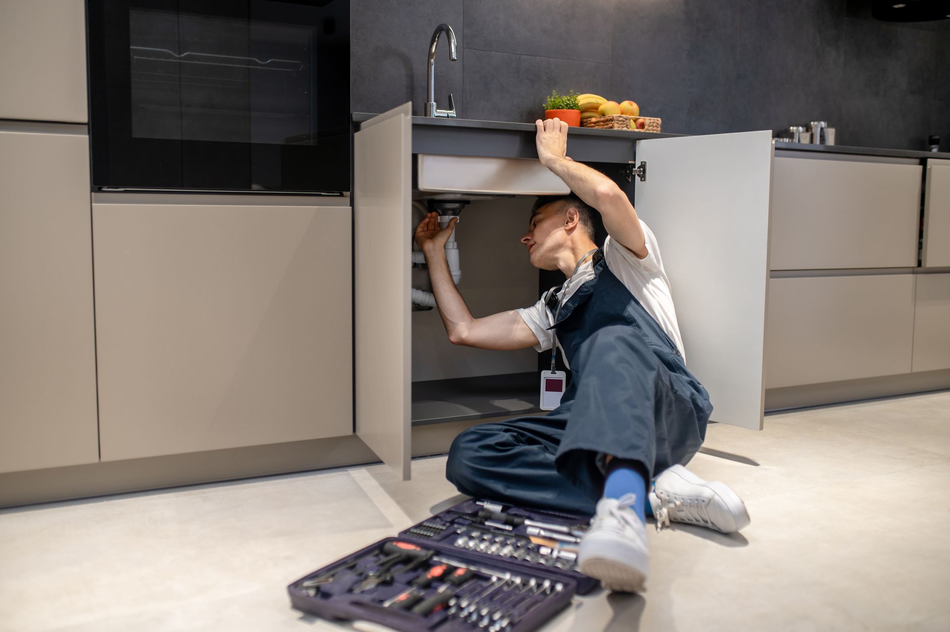 A person in blue overalls sits on a kitchen floor, repairing plumbing under the sink with a tool kit nearby.