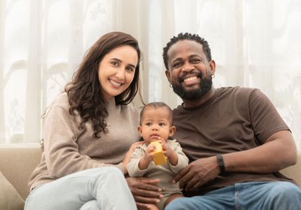 A smiling couple sits on a couch holding a baby, who is focused on a small, yellow toy, in a brightly lit room.
