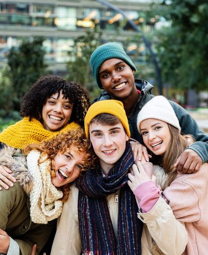 Five people wearing colorful knit hats and scarves huddle together outdoors, smiling warmly at the camera.