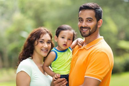 A smiling family poses for a photo outdoors with blurred greenery in the background.
