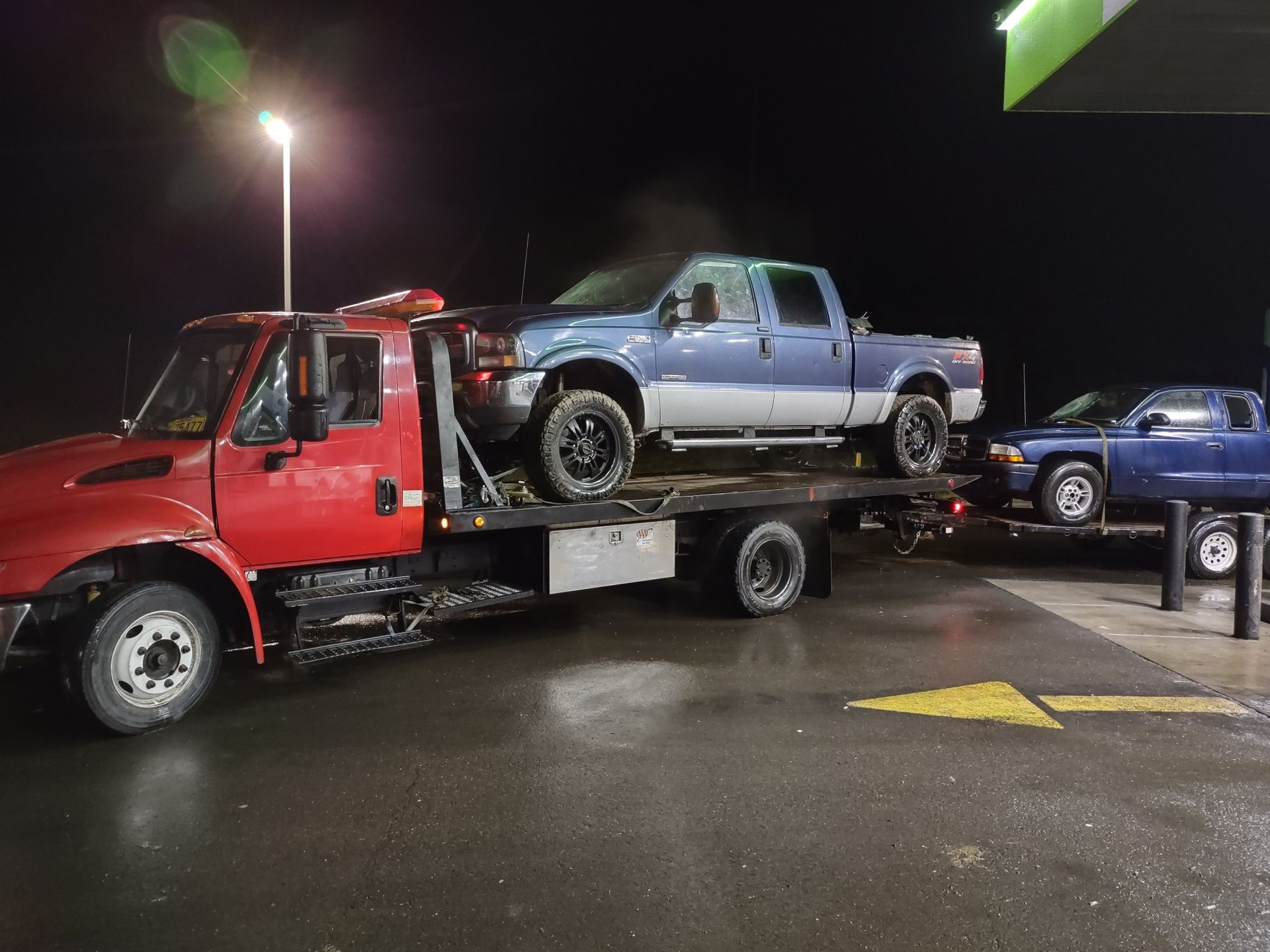 A red tow truck is towing a blue truck in a parking lot at night.