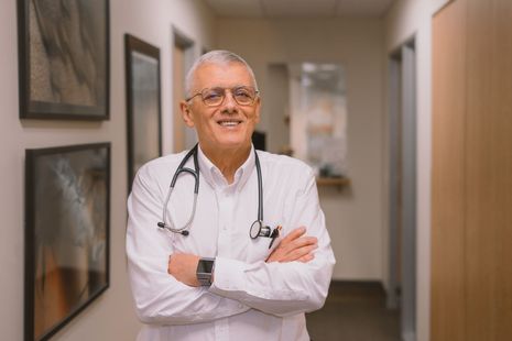 A doctor with a stethoscope around his neck is standing in a hallway with his arms crossed.