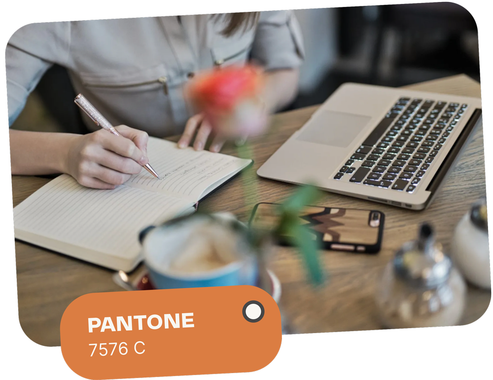 Person writing in a notebook next to a laptop and smartphone at a cafe table, with a Pantone color swatch: 7576 C.