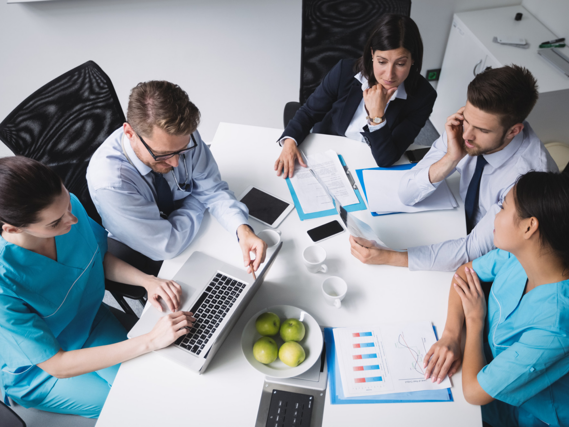 Five coworkers in a meeting around a white table, reviewing charts and laptops in an office.