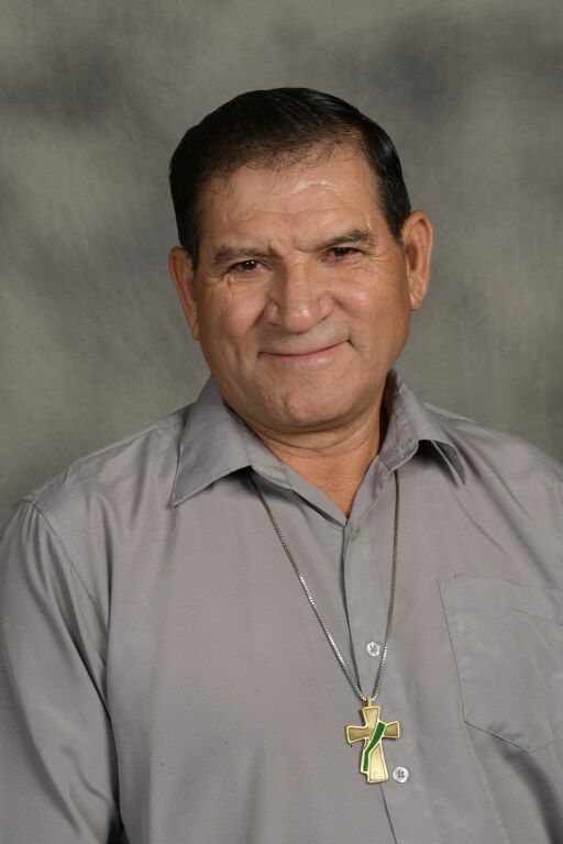 Man in gray shirt wearing a cross necklace, posed against a neutral studio backdrop.