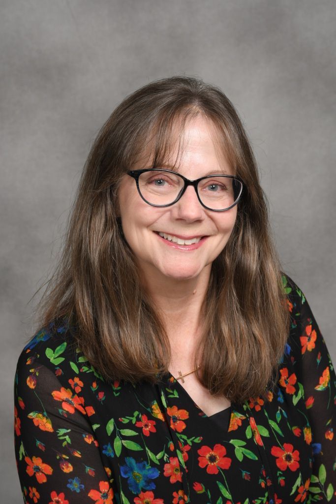 Smiling woman with glasses wearing a black floral blouse against a gray backdrop