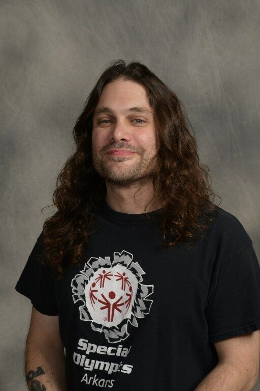 Person with long curly hair wearing a black Special Olympics T-shirt, standing against a gray backdrop.