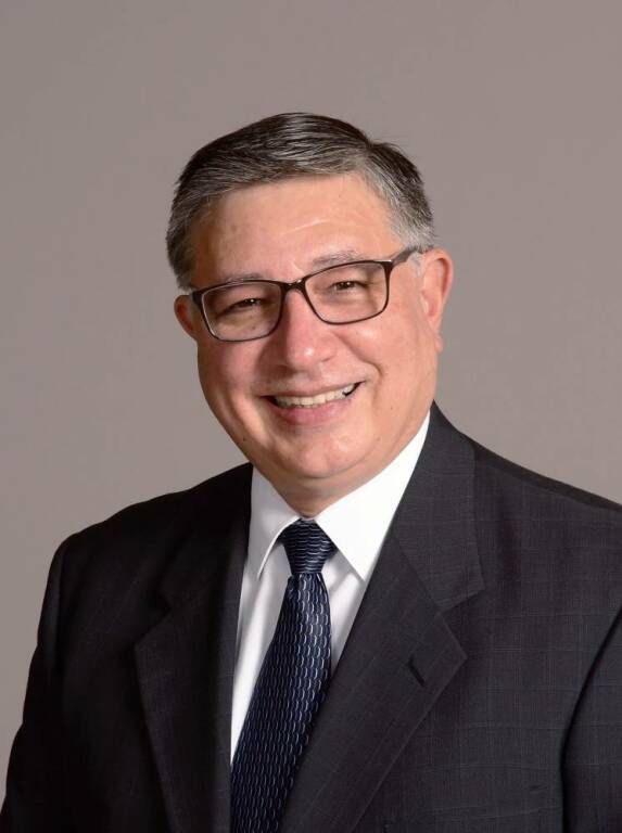 Smiling man in a dark suit, white shirt, and tie against a plain gray background
