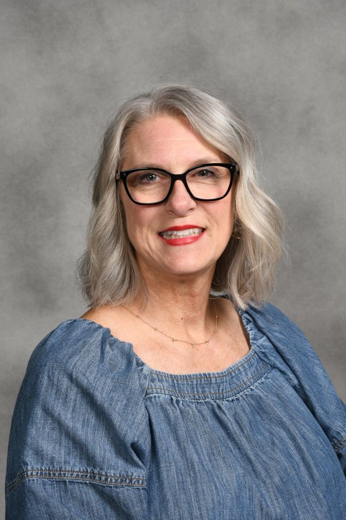 Portrait of a smiling woman with gray hair and glasses in a blue blouse against a gray backdrop