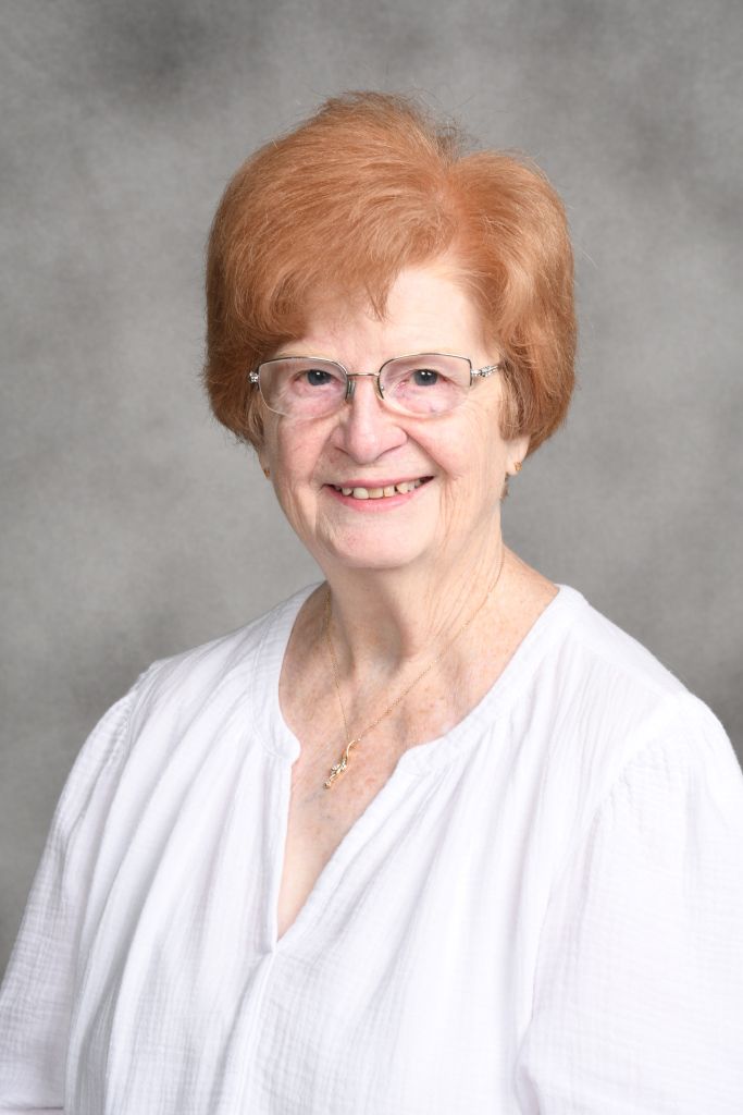Smiling woman with short red hair and glasses, wearing a white blouse against a gray studio backdrop