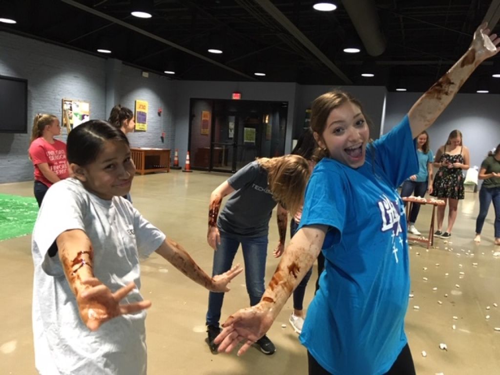 Children with painted arms pose playfully indoors, smiling and reaching toward the camera.