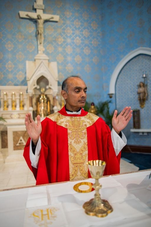 Priest in red vestments raises both hands during Mass at an altar with chalice and crucifix.