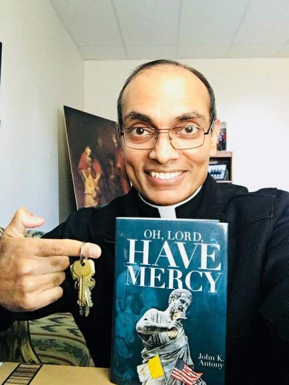 Smiling man holds a book titled “Oh, Lord, Have Mercy” and points at a small figurine beside it.