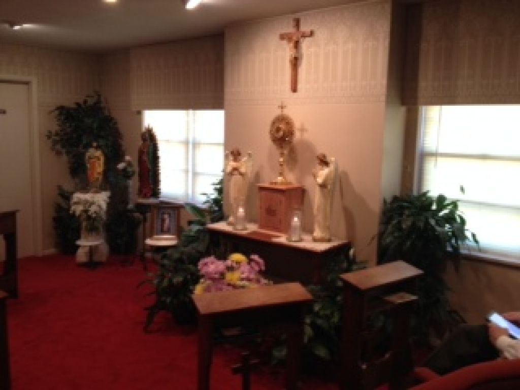 Small chapel interior with altar, crucifix, flowers, plants, and red carpeted floor