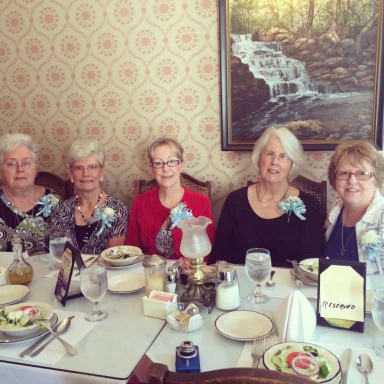Five women seated at a dining table in a decorated restaurant, with floral centerpieces and a waterfall painting behind them
