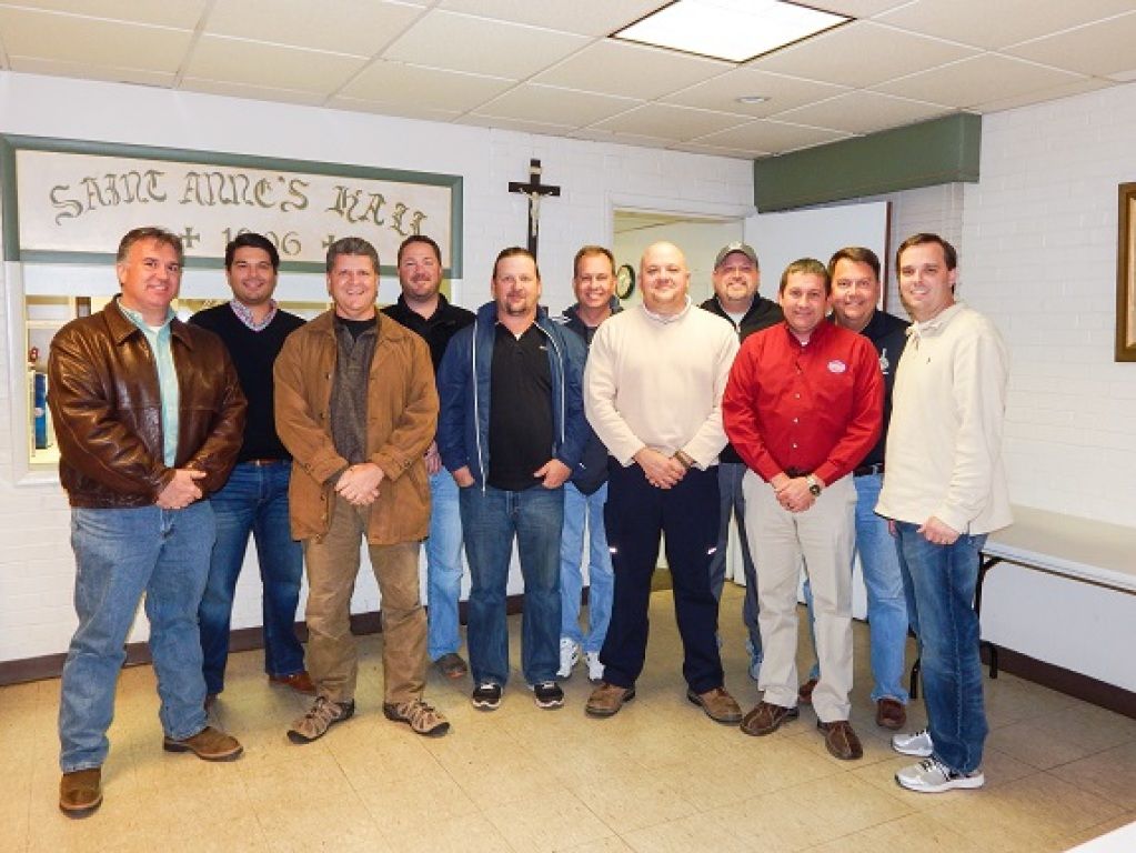 Nine men standing in a room with a banner and a cross, posing for a group photo