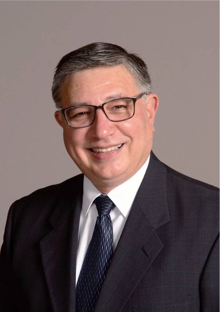 Smiling man in a dark suit, white shirt, and tie against a plain gray background