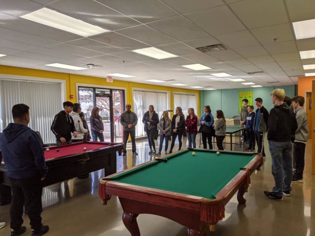 People standing around two pool tables in a recreation room, watching a game.