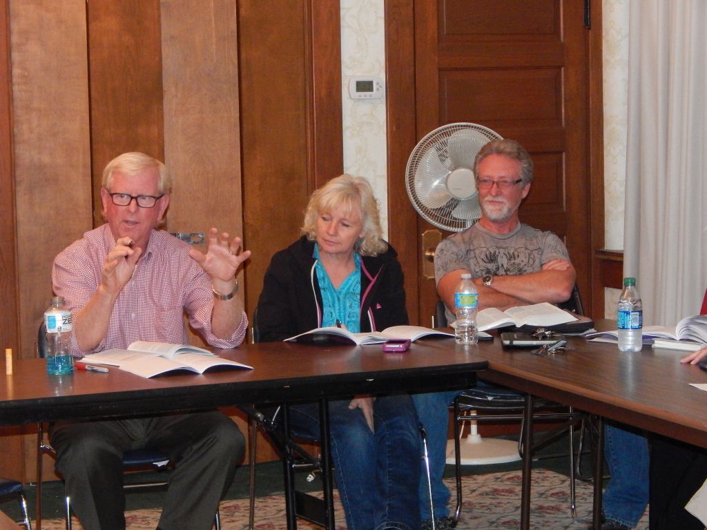 Three people seated at a conference table in a meeting room, one speaking while others listen